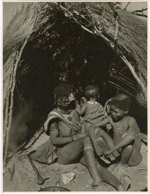 /Goishay, wife of ≠Gao, headman of Band 3, sitting with two of her children in front of a skerm; shows her scarification on her leg (print is a cropped image)