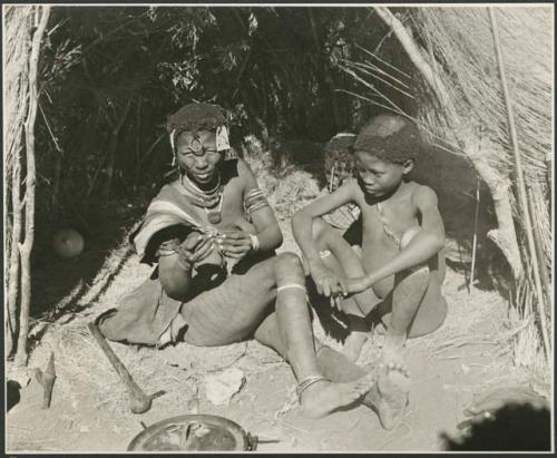 /Goishay, wife of ≠Gao, headman of Band 3, sitting with two of her children in front of a skerm; shows her scarification on her leg (print is a cropped image)