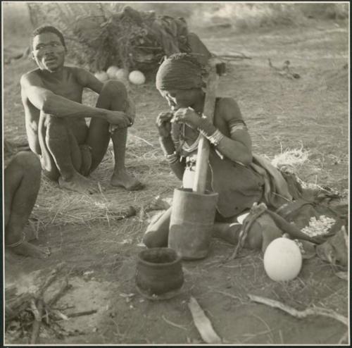 "Gao Medicine" sitting on grass, and Di!ai sitting by the fire and eating something she has pounded in the mortar (print is a cropped image)