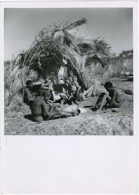 Gau of Band 2 sitting with his family in front of their skerm, including his son ≠Toma (by his first wife), his son !Naishi, Be (his first wife), Gau, Khuan//a (his second wife), their son /Qui, /Gunda (Khuan//a's son by a first husband), and ≠Gao (son of Be and Bau)