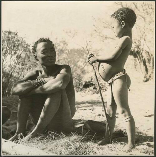 Bo sitting and looking at his daughter Be, who is standing next to him holding a stick  (print is a cropped image)