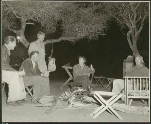 Two unidentified men sitting with John Marshall, Lorna Marshall and Laurence Marshall by a fire (print is a cropped image)