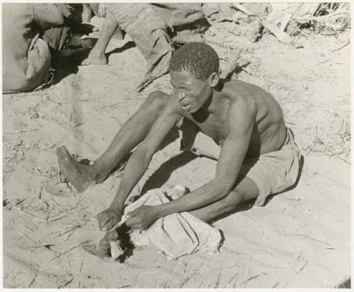 Man working on an animal skin, holding it with his foot (print is a cropped image)