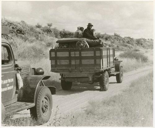 Expedition trucks on the road, two people sitting in the back of one truck (print is a cropped image)