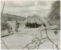 Woman standing outside of a hut (print is a cropped image)