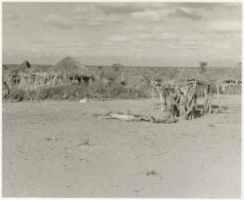 Two people standing in front of a hut, one person to the side in the clearing (print is a cropped image)