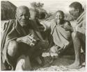 Elderly blind man sitting with three other people (print is a cropped image)