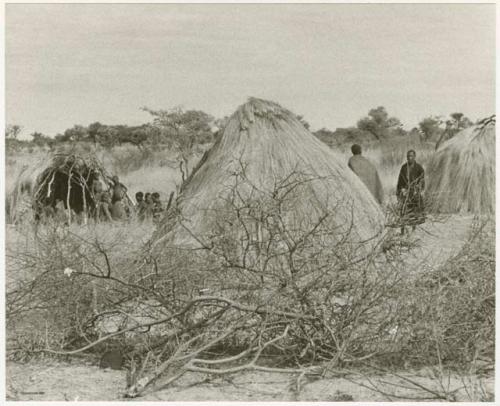 Kungwane huts in a group; two people standing amongst the huts and a group sitting in the background (print is a cropped image)