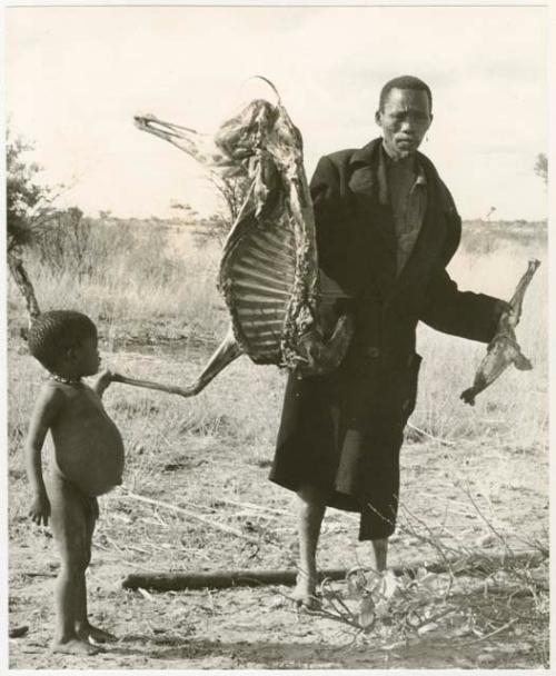 Man in a black overcoat holding up half an animal carcass (which the Marshalls gifted); a child standing next to him (print is a cropped image)