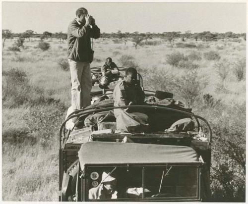 William Donnellan taking a picture on top of a truck; Simon Molamo and two other expedition members on the trucks (print is a cropped image)