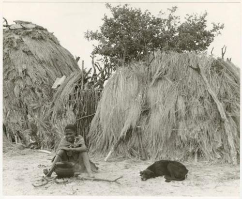 Boy and dog sitting before grass huts at Chukudu (print is a cropped image)