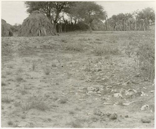 Ground and brush, shows a kraal with huts and a fence in the distance (print is a cropped image)