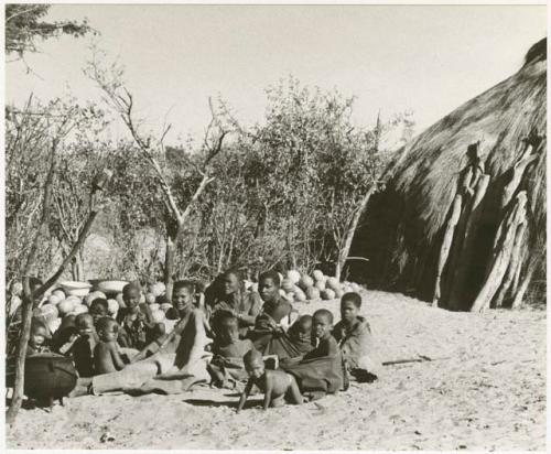 Group sitting beside a hut and a pile of tsama melons (print is a cropped image)