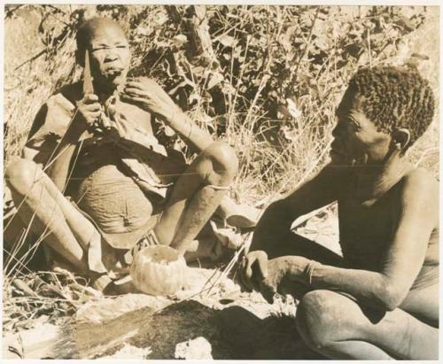 /Twikwe cutting meat at her lips with a knife, Oukwane in the foreground (print is a cropped image)