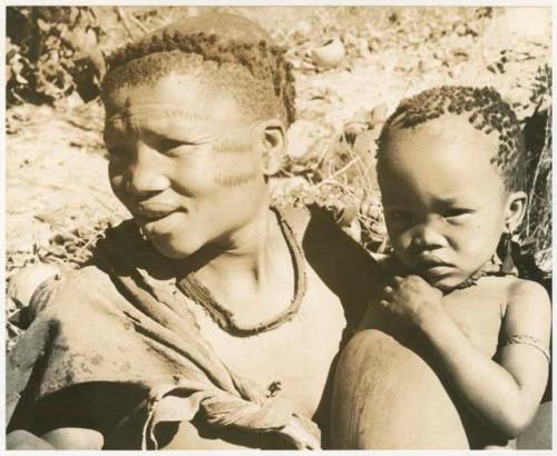 /Twikwe cutting meat at her lips with a knife, Oukwane in the foreground (print is a cropped image)