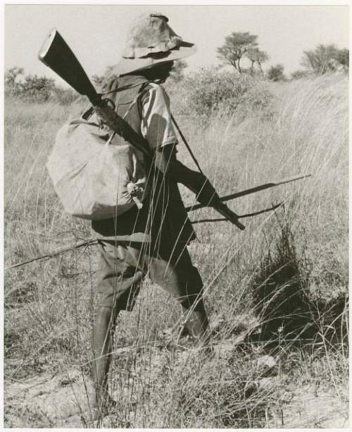 Headman walking, carrying a gun, a spear and a bundle, view from behind (print is a cropped image)