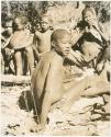 Group of people sitting, including Tsekue, N!hwakwe, /Gaiamakwe and !Gai, with /Twikwe eating in the background (print is a cropped image)