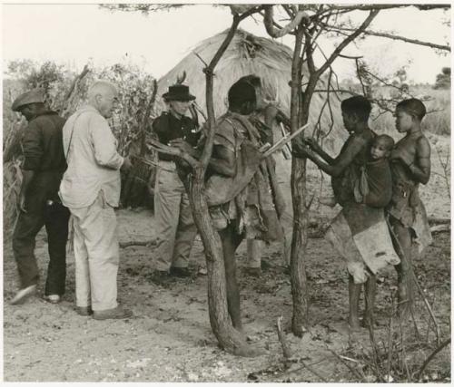 Ngwagetse, Genitsu (carrying a child), Rukan, and other man standing with Lorna Marshall, Laurence Marshall, and another expedition member (print is a cropped image)