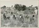 Group of people sitting with expedition members in the grass, John Marshall filming in the background