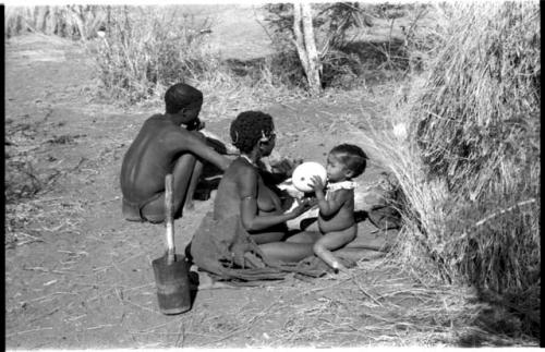 !Ungka (≠Toma's sister) with her daughter, Sa≠gai, sitting on her knees drinking from an ostrich eggshell; Tsamgao (Bau's husband) sitting beside her