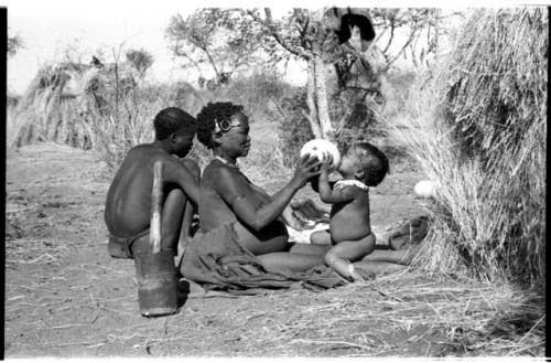 !Ungka (≠Toma's sister) with her daughter, Sa≠gai, sitting on her knees drinking from an ostrich egg shell; Tsamgao (Bau's husband) sitting beside her