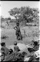 Group of Ju/'hoansi seated around a fire in the foreground; //Kushay ("Gao Helmet's" second wife) standing with her child on her shoulders in the background