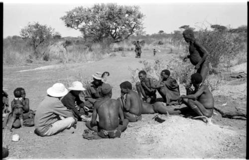 Laurence Marshall, Lorna Marshall, and Kernel Ledimo, wearing a hat with a feather, speaking with several men sitting on the ground including ≠Toma and ≠Gao (Khwo//o-/Gasa's husband); "Old Demi" standing to the right