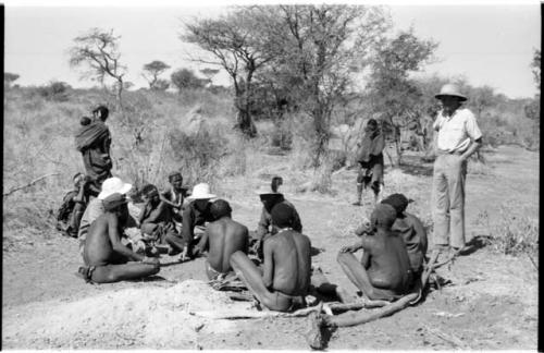 Laurence Marshall, Lorna Marshall, and Kernel Ledimo wearing a hat with a feather speaking with several men sitting on the ground including ≠Toma, ≠Gao (Khwo//o-/Gasa's husband), and "Old Demi"; Hans Ernst standing to the right and unidentified women in the background