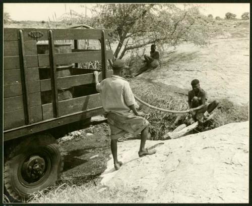 Man pumping water from a waterhole into water barrels on a truck and a man standing next to the truck; another man sitting in the background