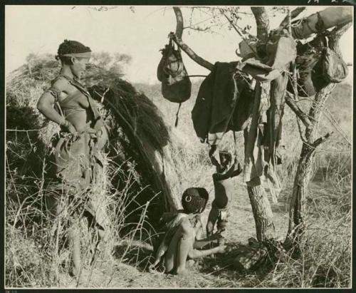 Woman and two children beside a skerm; belongings hang from a tree