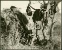 Woman and two children beside a skerm; belongings hang from a tree