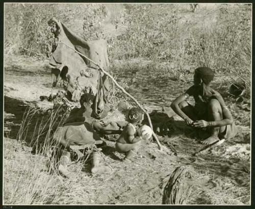 Man squatting, woman lying down, and child holding an ostrich eggshell, all around a fire; a kaross hangs on a bush and an unidentified object hangs from a branch stuck in the ground