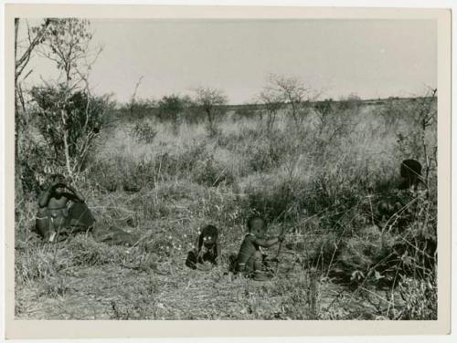 Two women sitting in grass and two children playing gathering veldkos, one holding a digging stick (print is a cropped image)