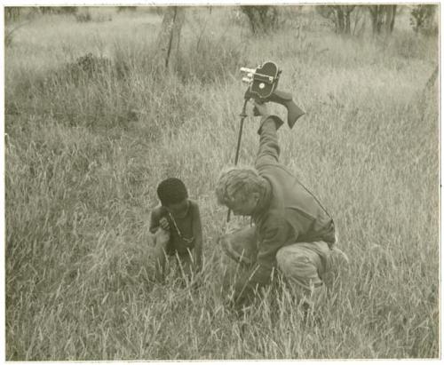 John Marshall crouching while holding a film camera and a Ju/'hoan boy crouching beside him