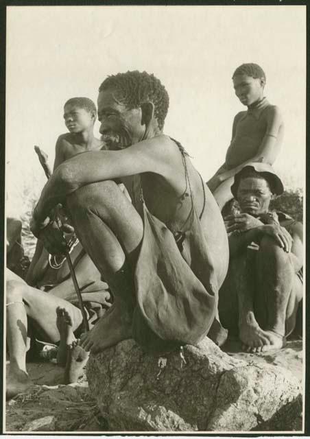 Ju/'hoan man crouching on a rock and other Ju/'hoansi behind him