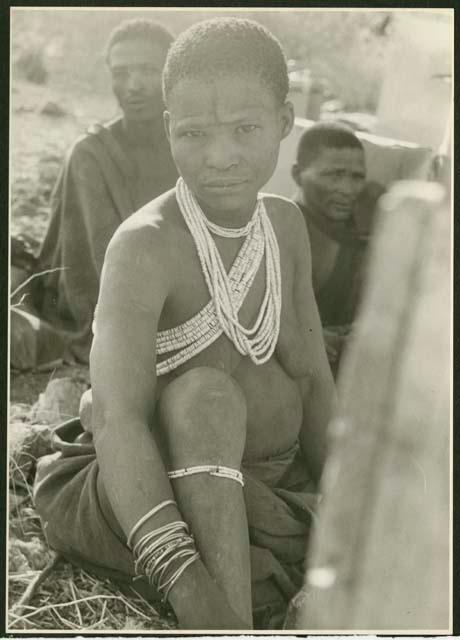 /Gasa (/Qui's wife) wearing an ostrich eggshell necklace; /Qui and another unidentified Ju/'hoan man in the background