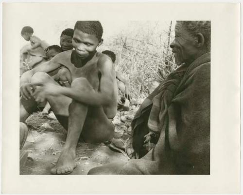 Elderly man and young man sitting; other people in the background (print is a cropped image)