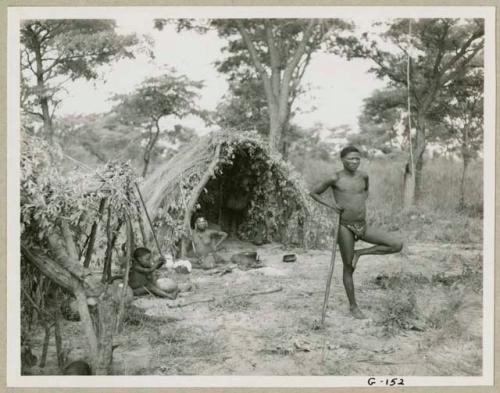 Man standing on one leg leaning against his stick, with woman and a child seated at skerm behind him