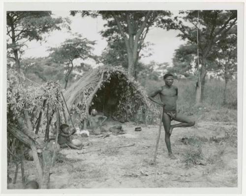 Man standing on one leg leaning against his stick, with woman and a child seated at skerm behind him