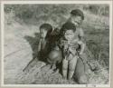 Woman sitting and holding a baby, another child beside her; baby is wearing a hunting talisman