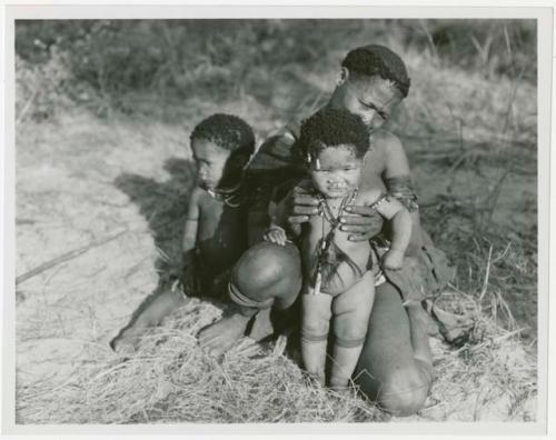 Woman sitting and holding a baby, another child beside her; baby is wearing a hunting talisman