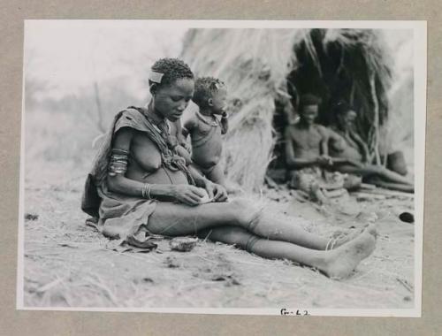 Woman seated, with Gau and one of his wives sitting in his skerm in the background



