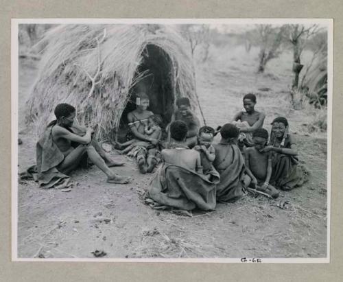 "/Qui Hunter" of Band 2 sitting in front of a skerm with his wife, /Naoka, holding her baby girl, /Khoa, and a group of people including Dam (husband of ≠Gisa, /Naoka's sister), Be (wife of Gau) with her son, and "Gao Scarf"