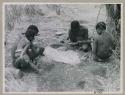 Group of men seated in front of a skerm, one taking his arrows out of a quiver
