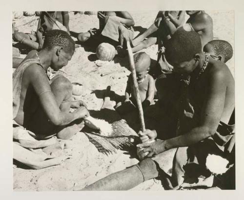 Woman sieving millet using a mat; woman (who is Kgalagari) opening a tsama melon with a digging stick; children and people in the background