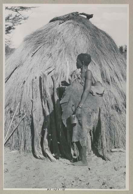 Woman with a kaross filled with something standing in front of a hut

