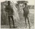 Three men standing, huts in background
