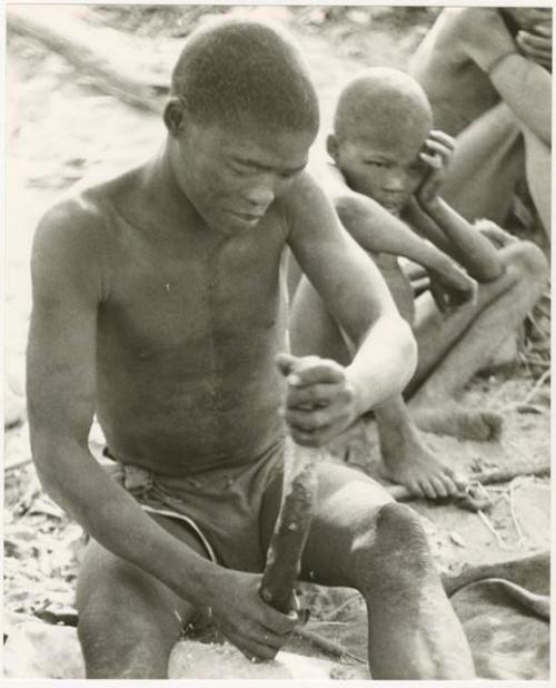 !Gai holding a piece of wood, two people in the background (print is a cropped image)