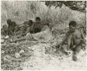 Oukwane holding cord with his toes, group sitting beside him (print is a cropped image)