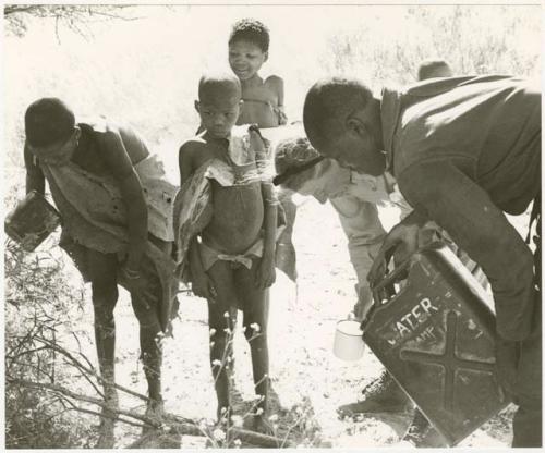 Simon Molamo and Lorna Marshall pouring water into a cup for three boys beside them (print is a cropped image)